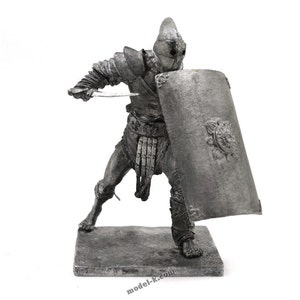 Puede incluir: Una figura de estaño de un gladiador romano sosteniendo una espada y un escudo. El gladiador lleva armadura y un casco. La figura está de pie sobre una pequeña base.