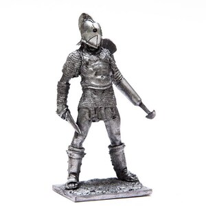 Puede incluir: Una figurita de metal plateada de un gladiador romano con armadura y sosteniendo una espada y un escudo.