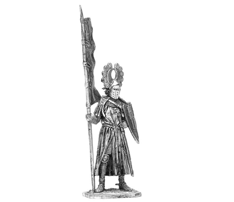 May include: A silver metal figurine of a knight in armour holding a flag and a shield. The knight is wearing a helmet with a crown and a chainmail tunic. The flag is red and white and has a design on it. The shield is round and has a design on it.