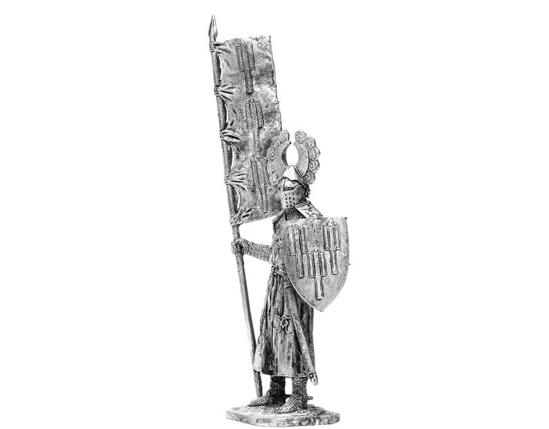 May include: A silver metal figurine of a medieval knight holding a flag and shield. The knight is wearing armour and a helmet with a crest. The flag is white with a red cross.