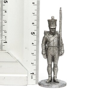 Może przedstawiać: Srebrna metalowa figurka żołnierza w wysokim kapeluszu trzymającego karabin. Figurka stoi na okrągłej podstawie i ma około 5 cm wysokości.