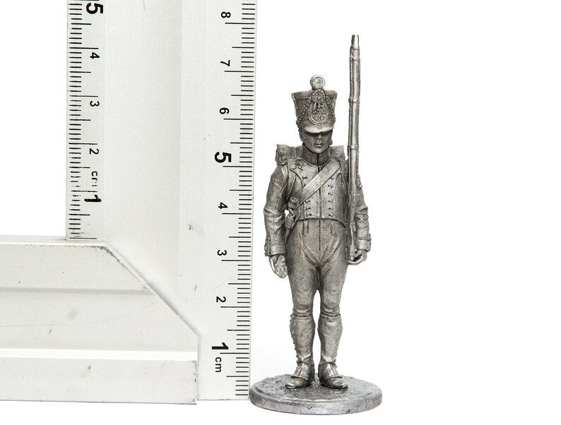 Może przedstawiać: Srebrna metalowa figurka żołnierza w wysokim kapeluszu trzymającego karabin. Figurka stoi na okrągłej podstawie i ma około 5 cm wysokości.
