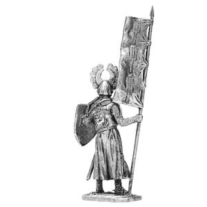 May include: A silver miniature figure of a medieval knight wearing armour and a helmet with a crest. The knight is holding a shield and a flag with a red and white design.