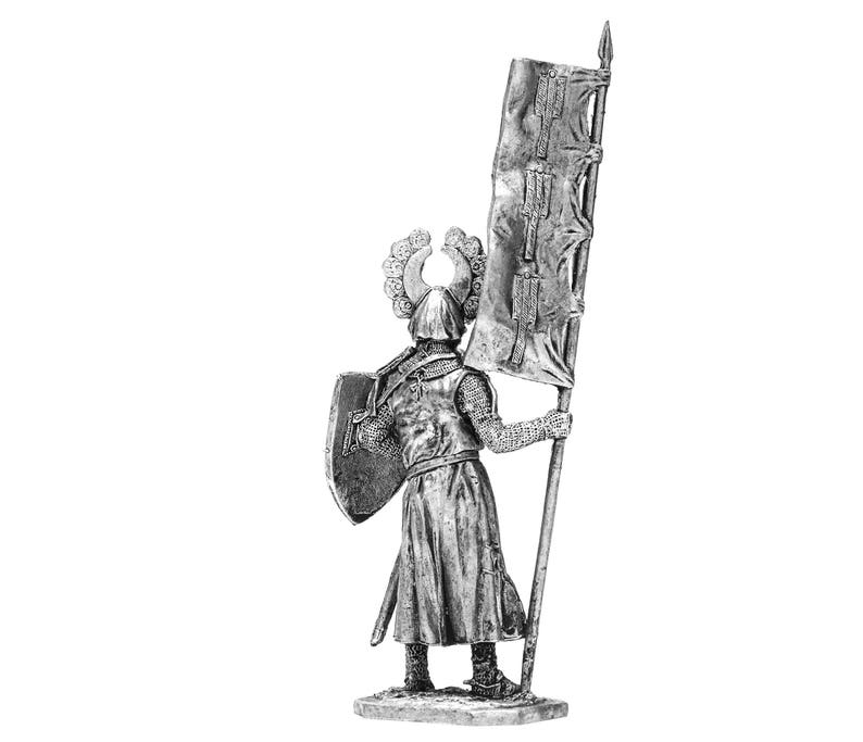 May include: A silver miniature figure of a medieval knight wearing armour and a helmet with a crest. The knight is holding a shield and a flag with a red and white design.