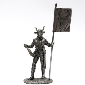 May include: A pewter figurine of a medieval soldier wearing a horned helmet and holding a flag with a bull's head design.