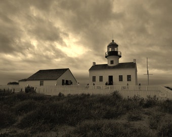 May include: A sepia-toned scene featuring a white lighthouse and an adjacent building enclosed by a white picket fence. The structures are set against a cloudy sky, with dark, low-lying vegetation in the foreground and a tall flagpole nearby.
