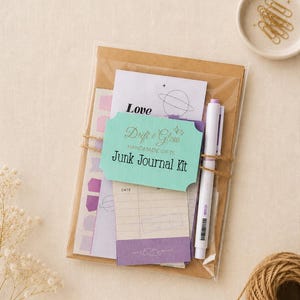 May include: A junk journal kit in a clear plastic sleeve, featuring a teal label with the text Drift & Glow Handmade Gifts Junk Journal Kit, a white pen with purple accents, and various paper stationery items including a card with the word Love.