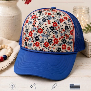 Op de afbeelding: Blauwe truckerpet met een bloemenpatroon op het voorpaneel. Het patroon heeft rode, blauwe en zwarte bloemen op een crèmekleurige achtergrond. De tekst "WILDFLOWER AMERICANA" staat erboven. De pet heeft een mesh achterkant en een gebogen klep.