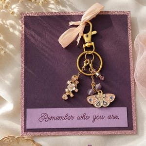 May include: A gold-colored keychain with a butterfly charm, flower charm, and purple beads. The keychain is attached to a purple card with the text "Remember who you are."