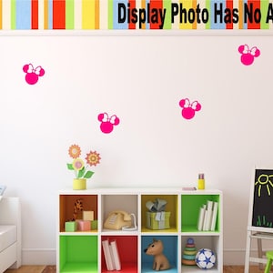 May include: Five pink Minnie Mouse head wall decals with white bows on a white wall. The decals are arranged in a staggered pattern. There is a white bookshelf with colorful shelves and a black chalkboard easel in the background.
