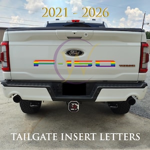 May include: A white Ford F-150 Tremor pickup truck tailgate featuring rainbow-colored F-150 insert letters. The text 2021 - 2026 appears at the top, and the text TAILGATE INSERT LETTERS appears at the bottom of the image.