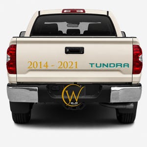 May include: The rear view of a beige pickup truck featuring the gold text 2014 - 2021 and the teal text TUNDRA on the tailgate. A black bumper with a central circular logo containing the letter W is visible below the tailgate and red taillights.