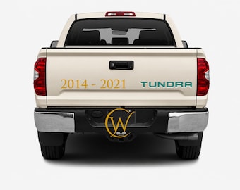 May include: The rear view of a beige pickup truck featuring the gold text 2014 - 2021 and the teal text TUNDRA on the tailgate. A black bumper with a central circular logo containing the letter W is visible below the tailgate and red taillights.