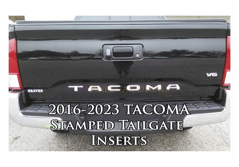 May include: Black 2016-2023 Toyota Tacoma tailgate with chrome trim and "TACOMA" stamped in silver lettering.