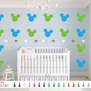 May include: A nursery room with a white crib and a blue wall decorated with 17+ color Mickey Mouse head wall decals. The decals are in various colors, including green, blue, and brown. The text "17+ COLORS" is at the bottom of the image.