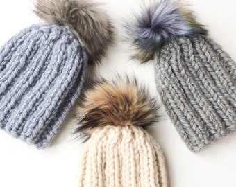 May include: Three knitted hats with faux fur pom poms. The hats are in gray, light gray, and white.