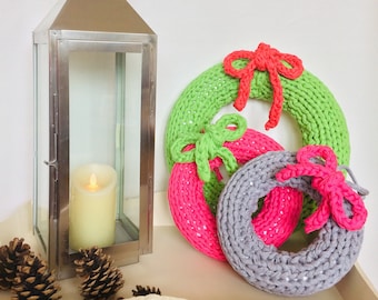 Könnte beinhalten: Drei gestrickte Kränze, einer grün, einer pink und einer grau, mit roten und grünen Schleifen, auf einer weißen Oberfläche mit einer silbernen Laterne und Tannenzapfen.