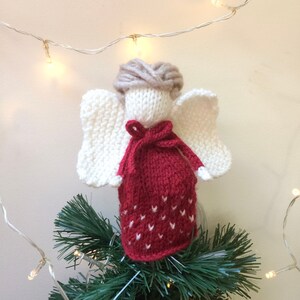 May include: A knitted angel ornament with white wings, a red dress with white hearts, and a brown yarn hairpiece. The angel is on top of a green Christmas tree.