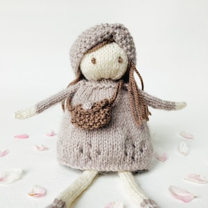 May include: A knitted doll with brown hair, a brown hat, and a brown bag. The doll is wearing a light brown dress and has white legs and feet. The doll is sitting on a white surface with pink flower petals scattered around it.