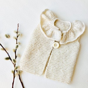 Può includere: Un gilet di maglia color crema per neonati con un colletto a volant e una chiusura con un solo bottone.