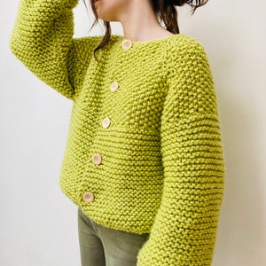 May include: A lime green chunky knit cardigan featuring a wide crew neckline, long sleeves with dropped shoulders, and five light-colored wooden buttons down the front. The garment is crafted with a thick, textured garter stitch pattern.