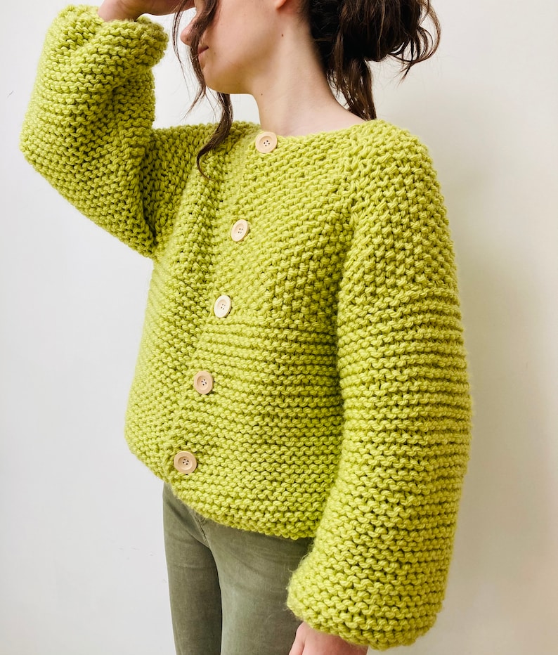May include: A lime green chunky knit cardigan featuring a wide crew neckline, long sleeves with dropped shoulders, and five light-colored wooden buttons down the front. The garment is crafted with a thick, textured garter stitch pattern.