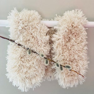 May include: A cream-colored, fluffy vest with a soft, textured appearance. The vest is displayed on a white rod, and a sprig of pussy willow adds a natural accent. The vest's design suggests a cozy, handmade aesthetic.