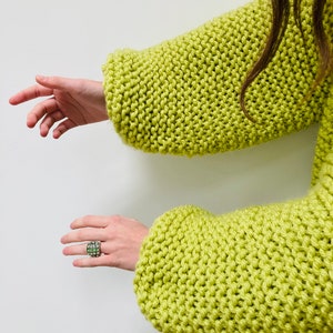 May include: A lime green chunky knit sweater with long sleeves and a thick, textured stitch pattern. A silver ring featuring a grid of small, light green square stones is worn on one finger, complementing the vibrant color of the knit garment.
