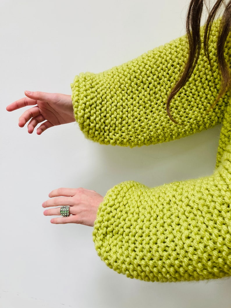 May include: A lime green chunky knit sweater with long sleeves and a thick, textured stitch pattern. A silver ring featuring a grid of small, light green square stones is worn on one finger, complementing the vibrant color of the knit garment.