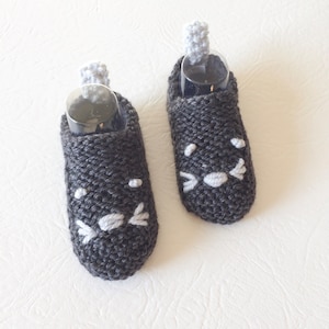 May include: A pair of gray knitted baby booties with white embroidered cat faces. The booties have a gray pom-pom on top.