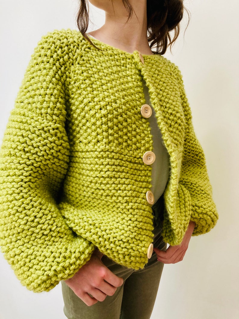 May include: A lime green chunky knit cardigan featuring a garter stitch texture, long sleeves with dropped shoulders, and a front button closure with four large, round, light-colored wooden buttons. The garment has a round neckline.