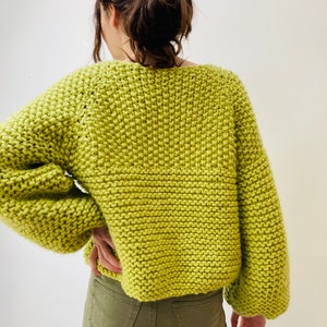 May include: A lime green chunky knit sweater with a wide neckline and long, loose sleeves. The garment features a thick, textured garter stitch pattern throughout, providing a bulky and soft appearance. The sweater is paired with olive green pants.