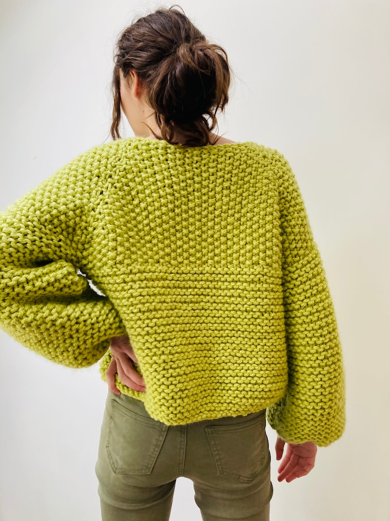 May include: A lime green chunky knit sweater with a wide neckline and long, loose sleeves. The garment features a thick, textured garter stitch pattern throughout, providing a bulky and soft appearance. The sweater is paired with olive green pants.