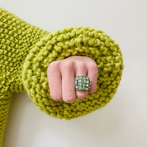 May include: A silver-toned ring featuring a square face with a grid pattern containing sixteen small, round, light green rhinestones. The ring is worn on a finger emerging from the cuff of a chunky, lime green, hand-knitted sweater.