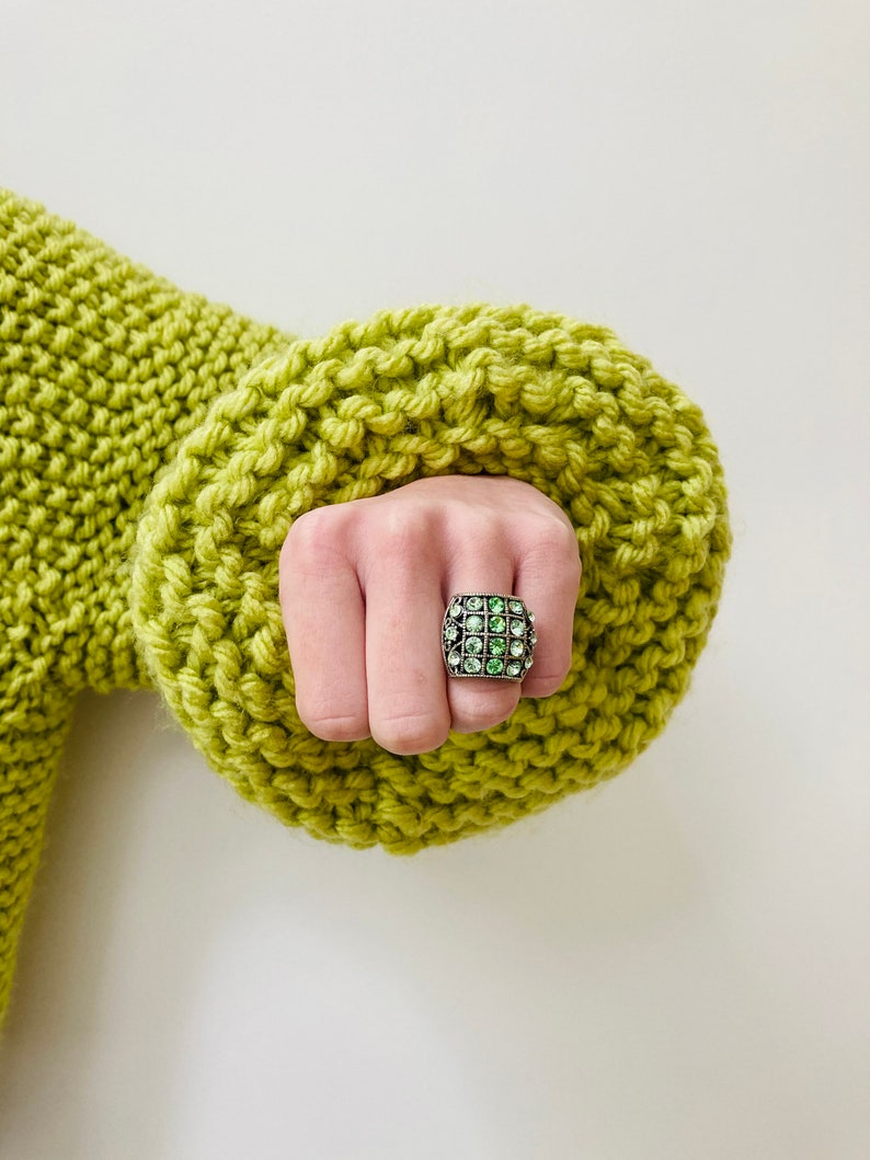May include: A silver-toned ring featuring a square face with a grid pattern containing sixteen small, round, light green rhinestones. The ring is worn on a finger emerging from the cuff of a chunky, lime green, hand-knitted sweater.