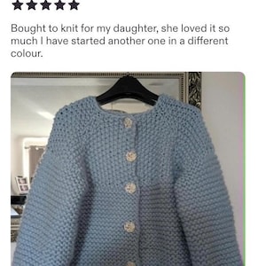 May include: A light blue chunky knit cardigan featuring a round neckline, long sleeves, and five round, textured white buttons down the front. The garment is displayed on a black hanger, showcasing a thick, textured yarn construction throughout.