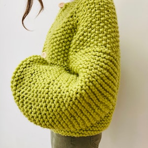 Op de afbeelding: Een felgroen, grofgebreid vest met houten knopen. De trui heeft een dikke, getextureerde steek en een relaxte pasvorm. De mouwen zijn volumineus en de stijl is oversized. Het vest wordt gedragen over een witte top en olijfgroene broek.