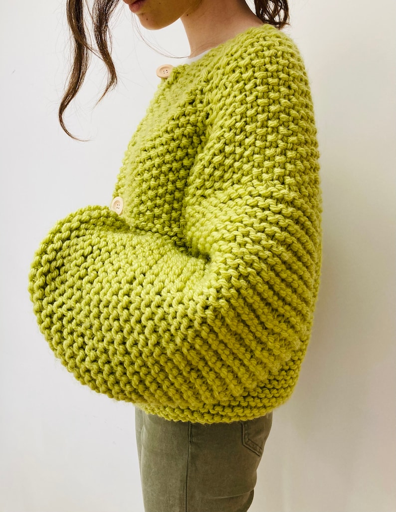 May include: A lime green chunky knit cardigan featuring a thick, textured stitch pattern, long sleeves, and two large, round, light-colored wooden buttons fastened along the front placket. The garment has a relaxed fit and a crew neckline.