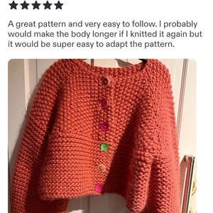 May include: A chunky knit cardigan in a solid coral color featuring a round neckline and long sleeves. The front closure is secured with five buttons in assorted colors, including light brown, pink, bright pink, green, and orange, arranged vertically.