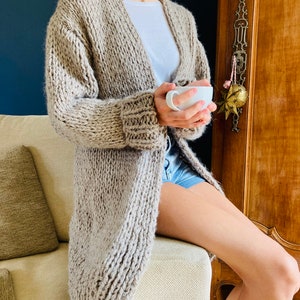May include: A long, open-front cardigan in a light beige color. The chunky knit sweater has a shawl collar and ribbed cuffs. The cardigan is worn over a white top and denim shorts. The person is holding a white mug.