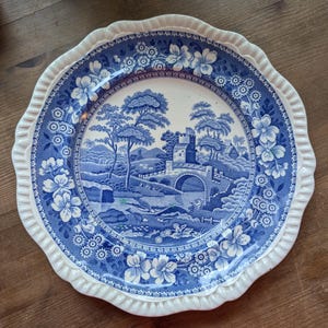 May include: A blue and white ceramic plate with a scalloped edge. The plate features a floral border and a central design of a bridge over a stream with a cottage in the background.