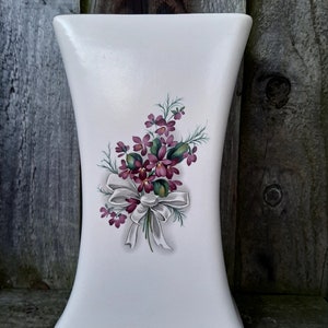 May include: White ceramic vase with a floral design featuring a bouquet of purple flowers with green leaves and a white ribbon.