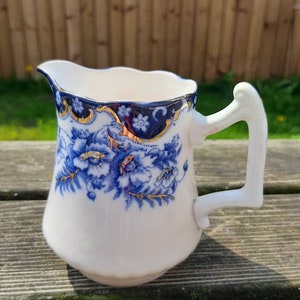 May include: A white ceramic milk jug with a blue and gold floral design. The jug has a scalloped rim and a handle.