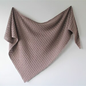 May include: A brown crocheted shawl with a lacy pattern. The shawl is draped over a white wall.