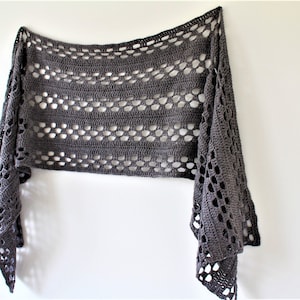 May include: A gray crochet shawl with a lacy pattern. The shawl is hanging on a white wall.