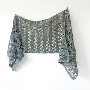 Puede incluir: Un chal de ganchillo de color verde salvia con un delicado patrón de calado. El chal está drapeado sobre una pared blanca.