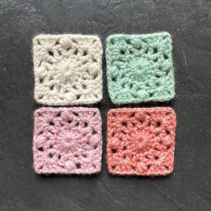 May include: Four handmade crochet squares in cream, mint green, light pink, and coral. Each square features a textured floral design. The squares are arranged on a dark gray surface, showcasing the vibrant colors and intricate details of the crochet work.