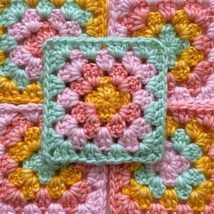 May include: A square crochet motif featuring a central yellow cluster surrounded by layers of pink, peach, and light green yarn. The pattern consists of repeating clusters of stitches forming a classic granny square design with a textured surface.