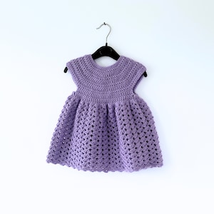May include: A purple crocheted baby dress with a scalloped hem and short sleeves. The dress is hanging on a black hanger against a white background.