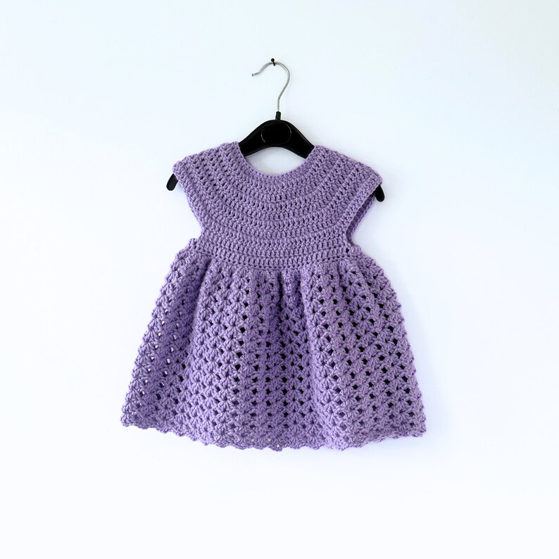 May include: A purple crocheted baby dress with a scalloped hem and short sleeves. The dress is hanging on a black hanger against a white background.
